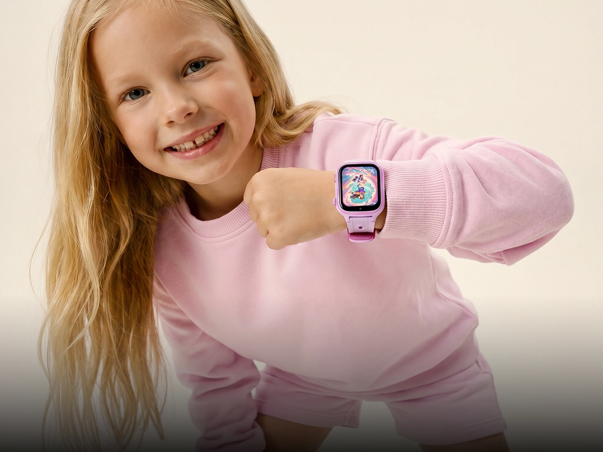 Uśmiechnięta dziewczynka w różowej bluzie pokazuje smartwatch dziecięcy Besti z różowym paskiem i kolorową tarczą.