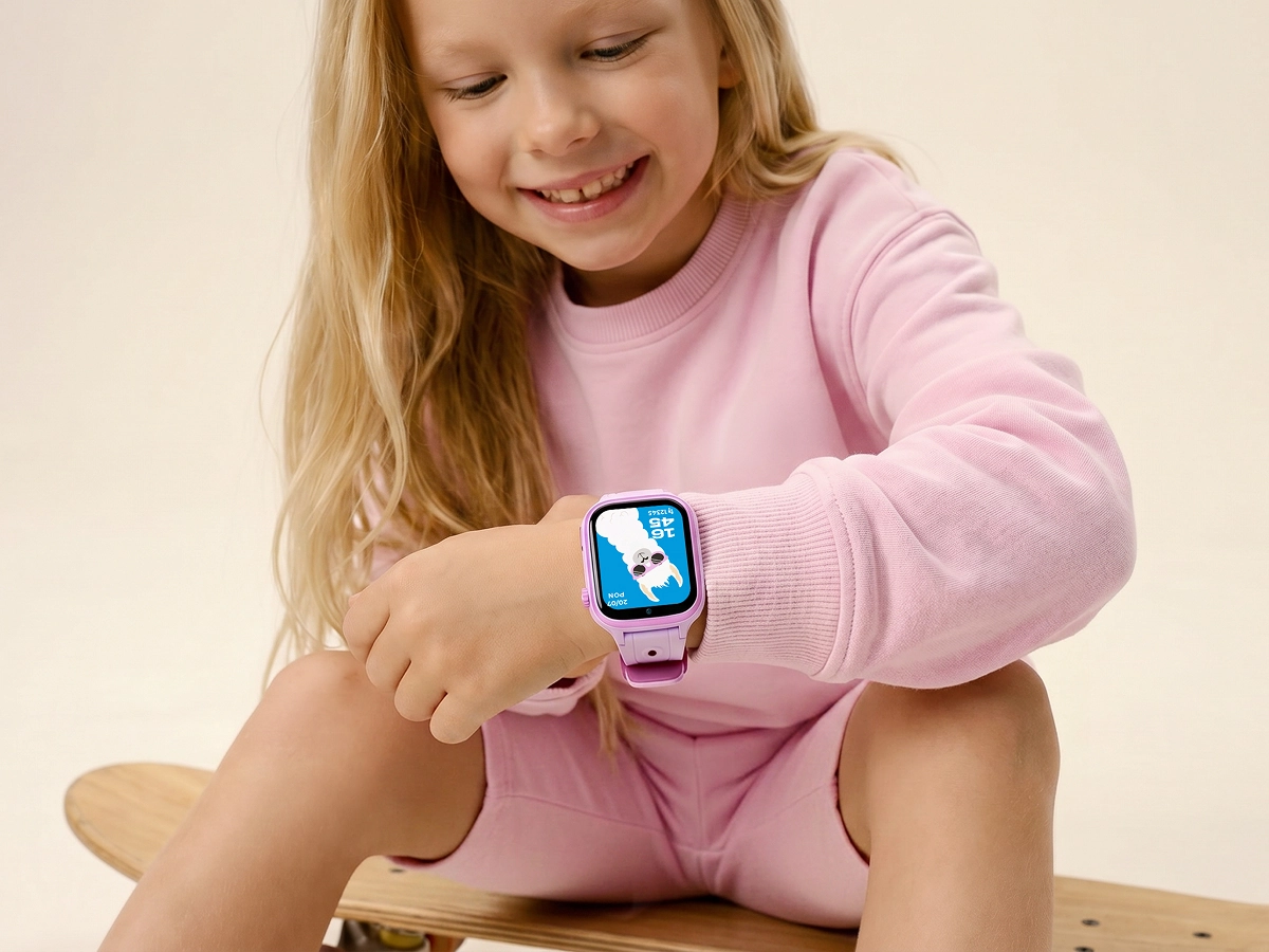 Uśmiechnięta dziewczynka siedzi na deskorolce i patrzy na różowy smartwatch dziecięcy Besti.
